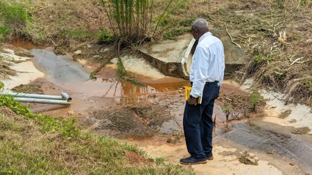 Robert Bullard wears a white shirt and stands over a flooded drainage area that is surrounded by mud