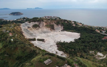 Aerial view of the Solfatara crater surrounded by green landscape and villages by the sea.