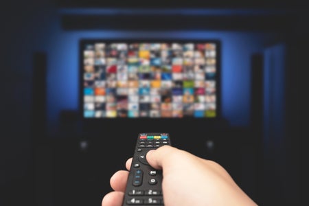 Person with remote control in hand pointing at TV screen in a dark room.