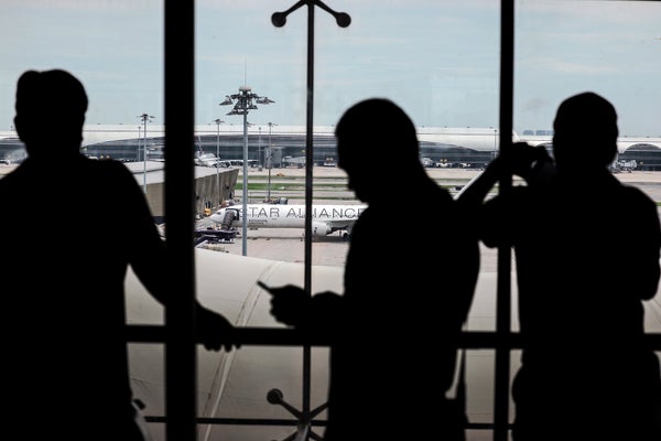 Photograph looking outwards from an airport window to a parked Signapore Airlines flight on the tarmac as passengers seen in silhouette stand in front of the window taking photos, looking at the plane, and using their phones