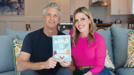 Penn (black T-shirt) and Kim (pink sweater) Holderness sitting on a couch between pillows holding up their book titled: "ADHD is Awesome."